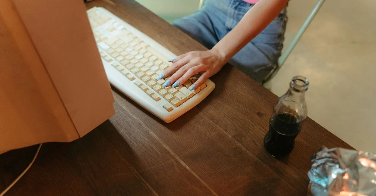Une personne avec des ongles manucurés bleus utilisant un clavier d'ordinateur sur une table en bois avec des collations et une boisson.